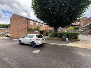 Une rue résidentielle avec une voiture blanche garée sur le bord, un bâtiment en briques et un arbre.