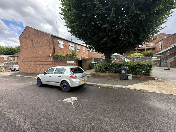 A residential street with a white car parked on the side, a brick building, and a tree.