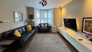 Ein modernes Wohnzimmer mit einer schwarzen Ledersofa, einem kleineren Sofa, einem großen Flachbildfernseher an der Wand und einer raumhohen Unterhaltungsanlage.