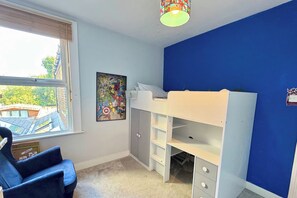 A child's bedroom with a bunk bed, a desk, a chair, a window, and a colorful wall decoration.