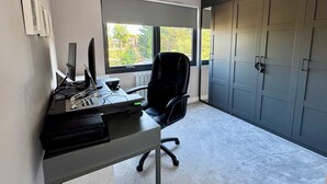 Ein moderner Arbeitsraum mit Schreibtisch, Computer und Stuhl, einem großen Fenster mit Blick auf Grünflächen und einem grauen Kleiderschrank.