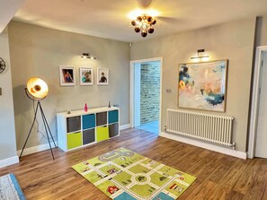 A room with a wooden floor, a colorful play mat, a white console table with storage, a floor lamp, framed photos on the wall, a radiator, and a large abstract painting.
