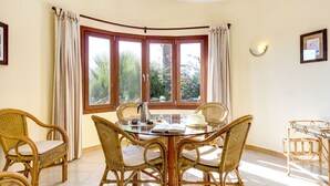A dining area with a round table, wicker chairs, and a view of greenery through large windows. - Villa Los Leones - Three Bedroom Villa, Sleeps 6 (Alayor)