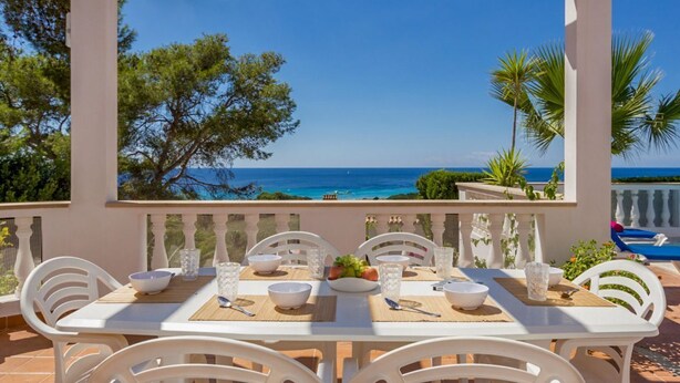 A terrace with a table set for dining, overlooking a coastal view with trees and a clear sky.