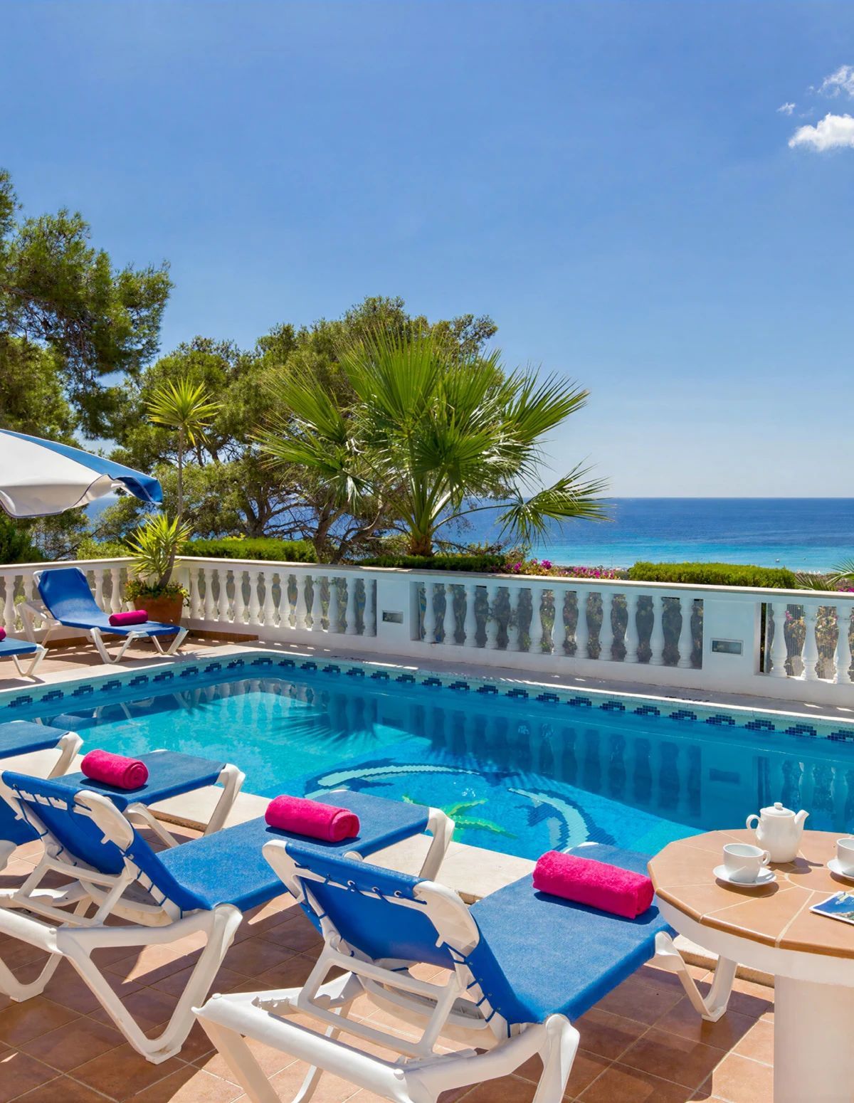 A pool area with lounge chairs and a table, surrounded by a white balustrade, overlooking a clear blue sea.