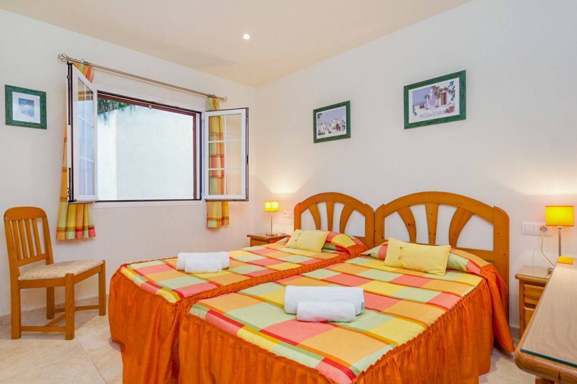 Un lit double avec une couverture colorée, une chaise en bois, une petite table et une fenêtre avec des rideaux.