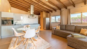 Una cocina moderna con una mesa de comedor de vidrio, sillas blancas y un horno empotrado. Hay un sofá gris y una ventana con vistas a zonas verdes.