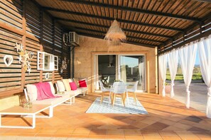 A covered patio with a wooden ceiling, a sofa with cushions, a table with chairs, and a hanging light fixture.