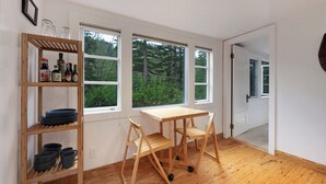 A room with a wooden floor, a table with chairs, a shelf with bottles, and three windows with a view of greenery.