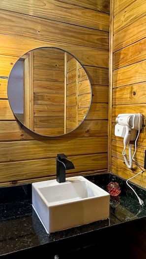 A wooden interior with a white sink, a round mirror, and a hairdryer on the wall. - Casa de Praia (Buzios)