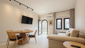 Un salon moderne avec un canapé beige, une table de salle à manger ronde entourée de chaises, un téléviseur à écran plat fixé au mur et de grandes fenêtres avec des rideaux.