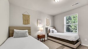 A bedroom with two beds, a window with blinds, a bedside table with a lamp, and wall art.