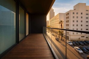 Un balcón con piso de madera, barandas de vidrio y vista a una calle urbana con automóviles y edificios.