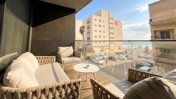 A balcony with wicker furniture, a small table, and a view of the street and buildings. - #SeaView Beach Front 3BR TLV  | By SweetyGuest (Tel Aviv-Yafo)
