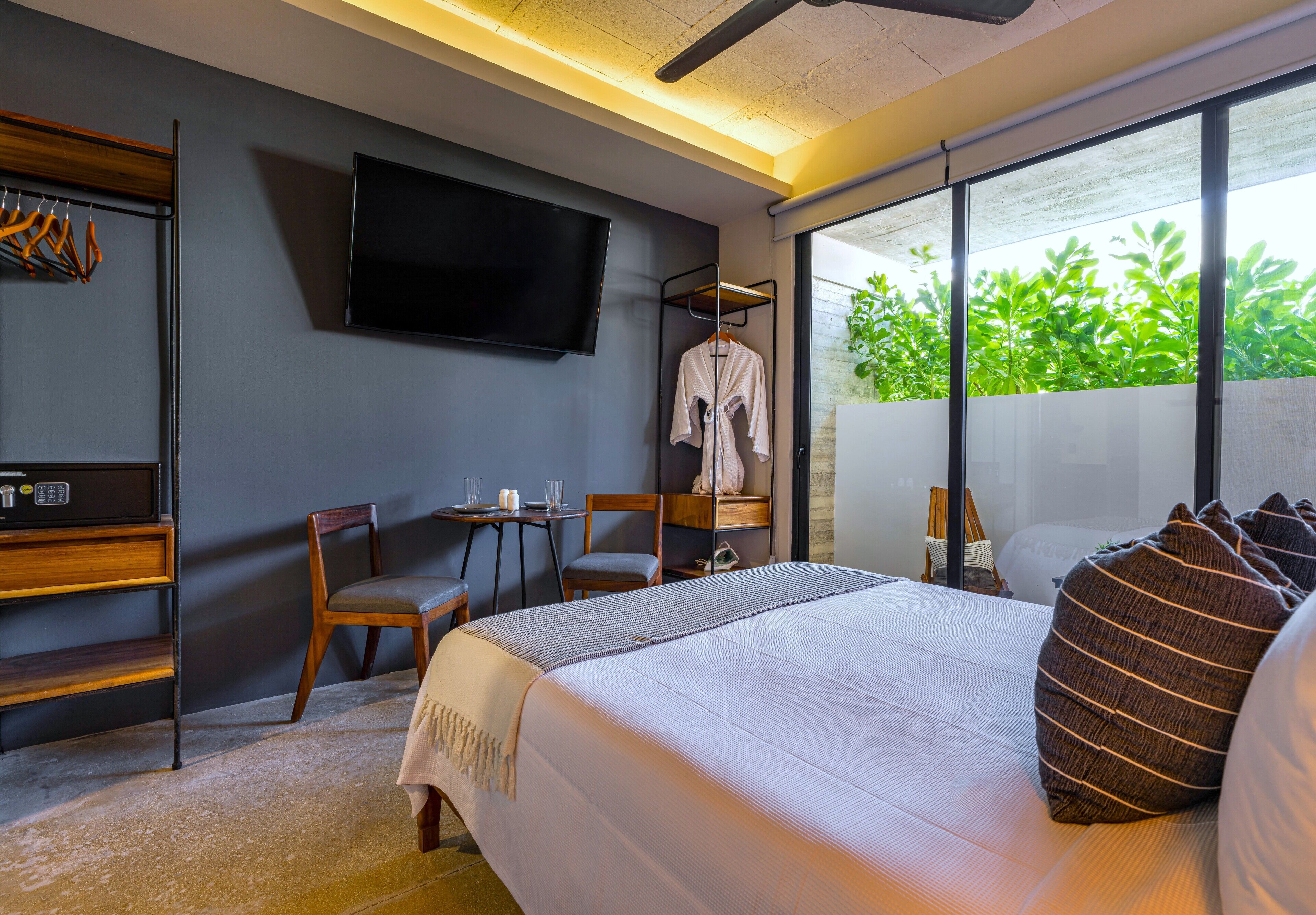 Una habitación de hotel moderna con una cama grande, un televisor en la pared, un espacio de descanso con una mesa y sillas, y vistas a zonas verdes por la ventana.