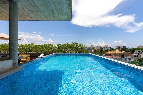 A rooftop swimming pool with clear blue water, surrounded by lounge chairs and a view of the city skyline.