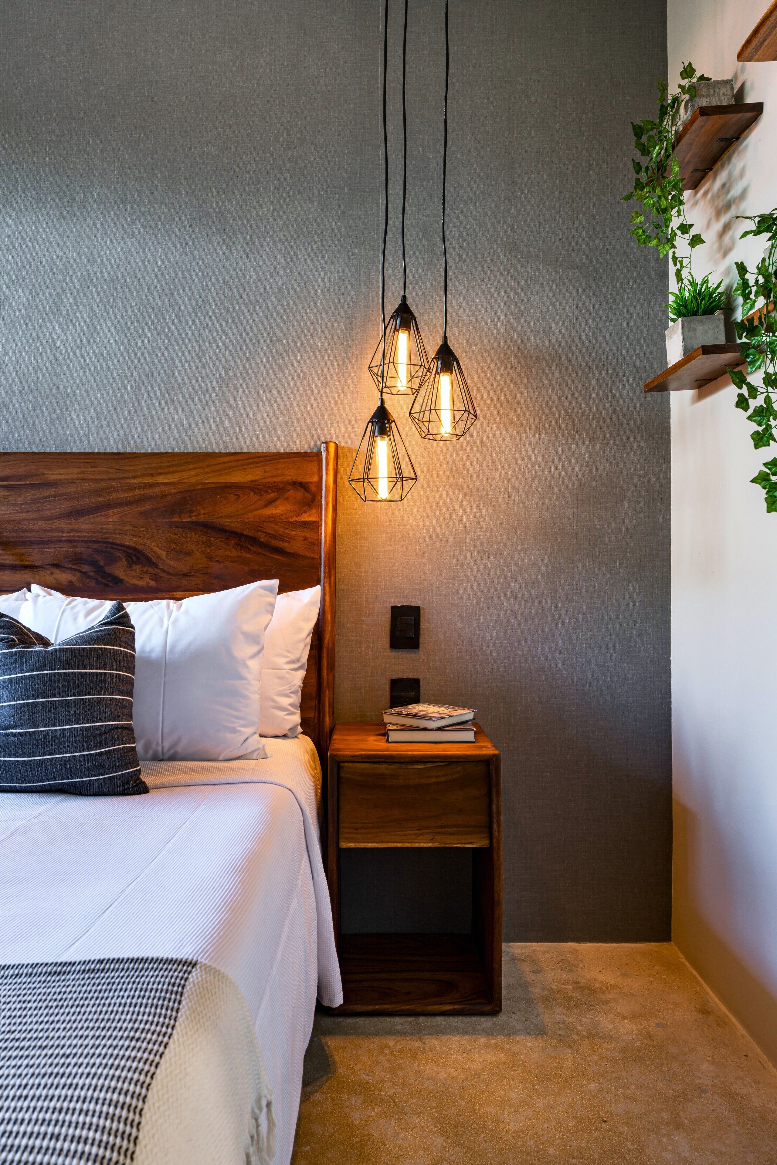 A neatly made bed with white linens and a striped pillow, a wooden nightstand with a book and a lamp, and a wall-mounted shelf with plants.