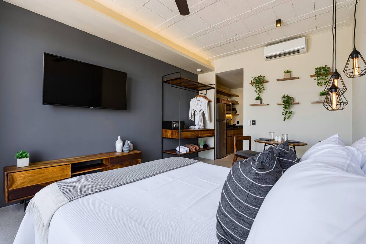 A modern hotel room with a large bed, a TV mounted on the wall, a wooden console table, a seating area with a table and chairs, and a kitchen area visible through an open door.