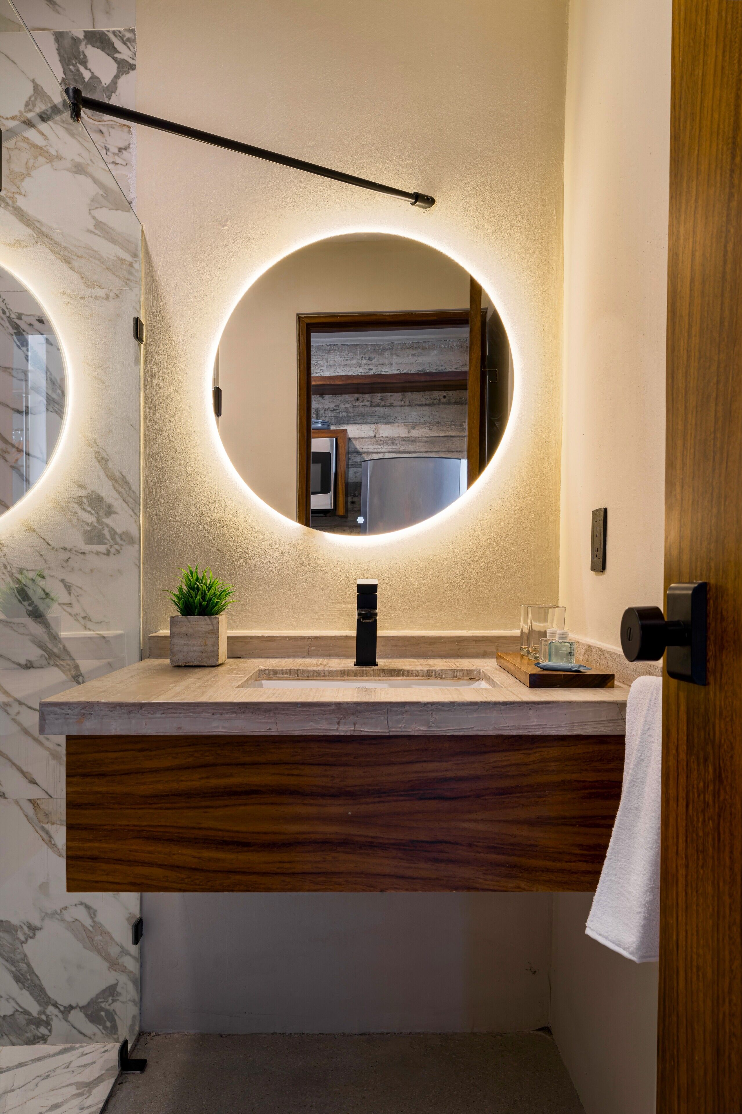 Une salle de bain moderne avec un mur en marbre, un meuble-lavabo en bois, un grand miroir rond doté d’un éclairage indirect et une petite plante en pot.
