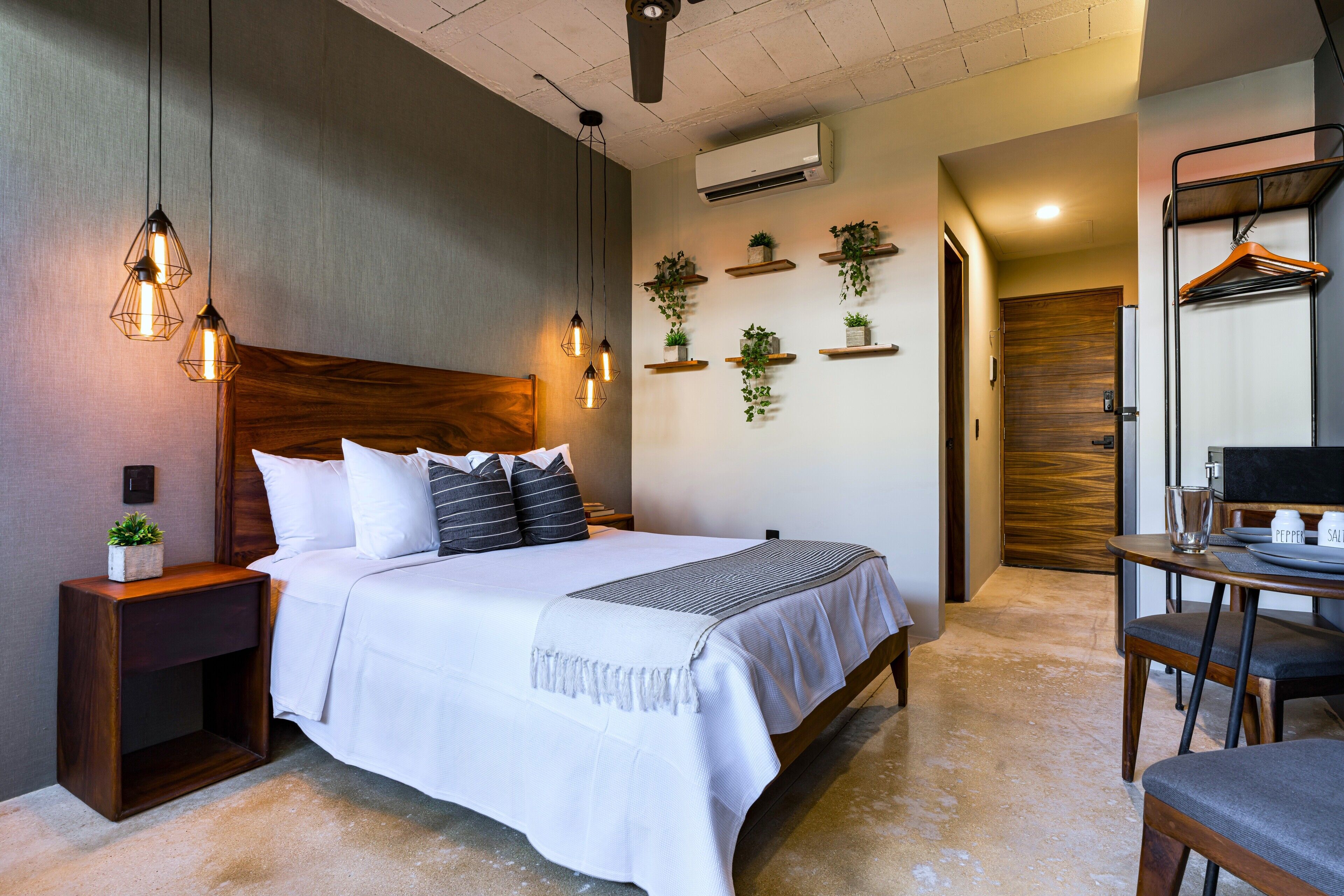 A neatly arranged bedroom with a wooden headboard, a bed with white bedding, bedside tables with lamps, a small plant, and a wall-mounted shelf with potted plants.