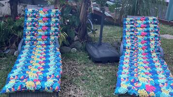 Two outdoor lounge chairs with colorful tropical leaf-patterned cushions, positioned under a large banana tree in a garden setting.