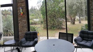 A patio area with a round table and two chairs, large windows with a view of a garden, and a brick wall. - Large villa with 11x5m pool in 7700 M2 grounds. Veranda and multiple terraces. (Montescot)