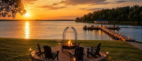 A lakeside patio with a fire pit, chairs, and a dock extending into the water.