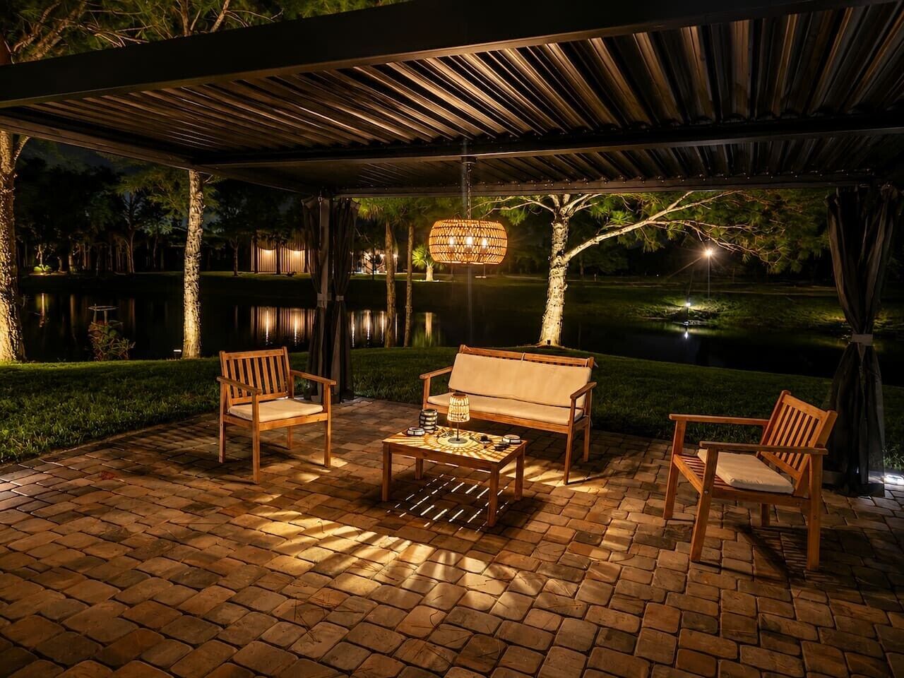 Een terras buiten met een houten tafel en stoelen, een hanglamp en uitzicht op een vijver.