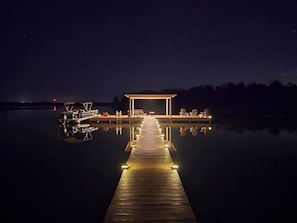 Eine hölzerne Anlegestelle führt zu einem überdachten Bereich mit Sitzgelegenheiten über dem Wasser, bei Nacht.