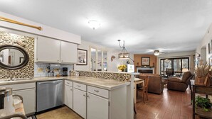 A modern kitchen with white cabinets, a dishwasher, and a tiled backsplash. There is a dining area with a table and chairs, and a living room with a sofa and a coffee table. - Water's Edge Hideaway – 6ppl (Branson)