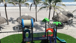 Une aire de jeux avec des balançoires, des toboggans et des structures d’escalade, entourée de palmiers et de parasols de plage, le tout sur une plage de sable.