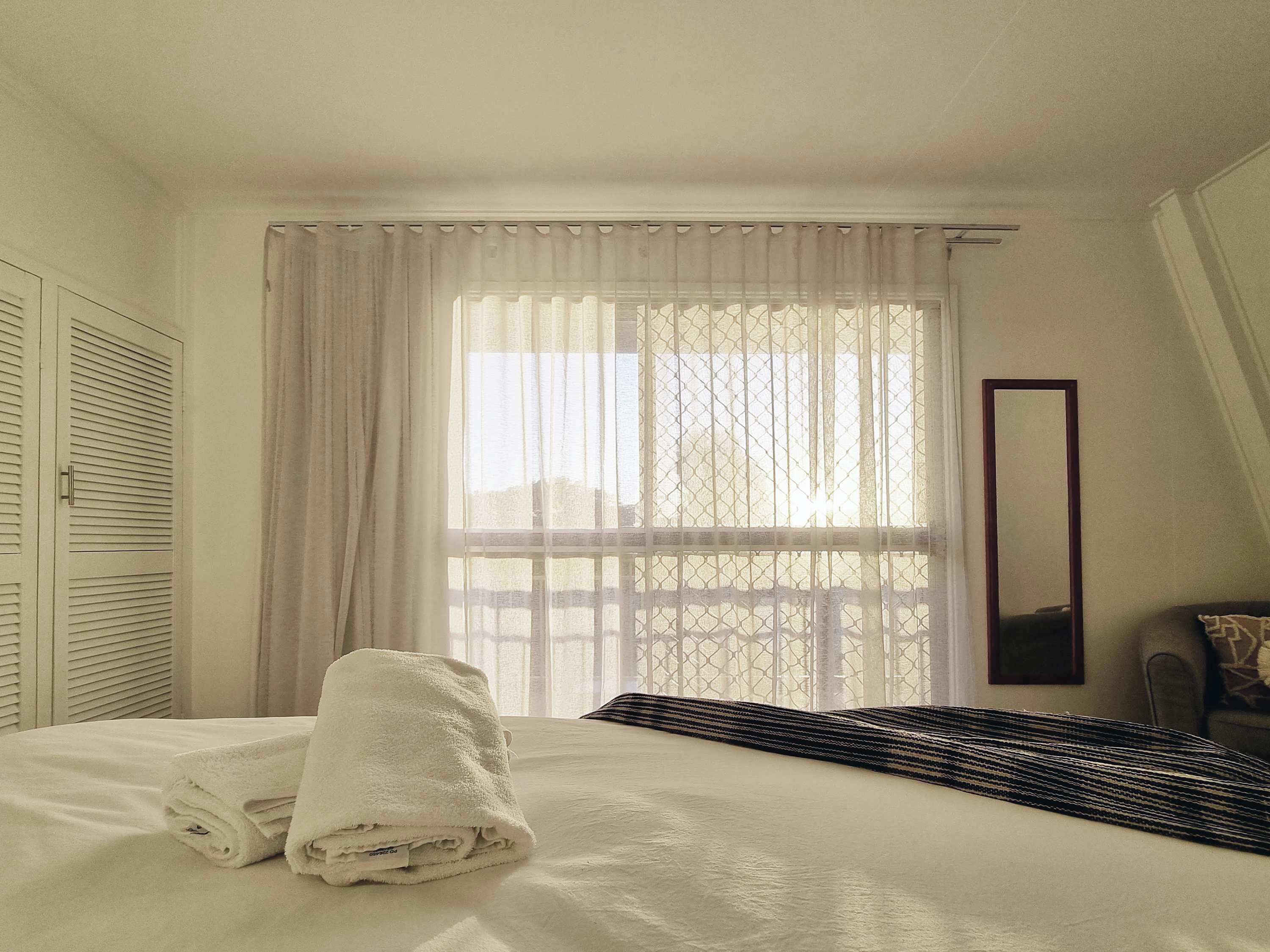 A hotel room with a bed, a window with sheer curtains, a mirror, and a folded towel on the bed.