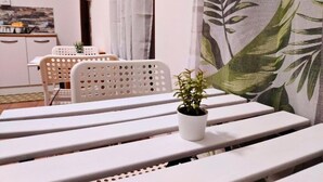 Een eetgedeelte met witte plastic stoelen en banken, een kleine plant in een pot op een bank, en een keuken in de achtergrond.