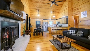 A cozy log cabin interior with a fireplace, dining area, kitchen, and a living room with a sofa and coffee table.