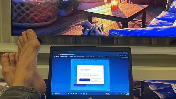 Un ordinateur portable posé sur un bureau, avec une fenêtre demandant un code de sécurité, un téléviseur diffusant une scène de plage, et une personne allongée, les pieds en l’air.