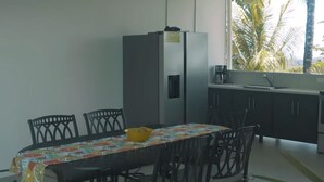 Un coin repas comprenant une table, des chaises, un réfrigérateur et une fenêtre donnant sur des palmiers.