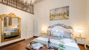 A bedroom with a large ornate mirror, a bed with a white headboard, two bedside lamps, a painting on the wall, and a wooden table with a bottle and glasses. - Bright and Central Apartment in Lucca (Lucca)