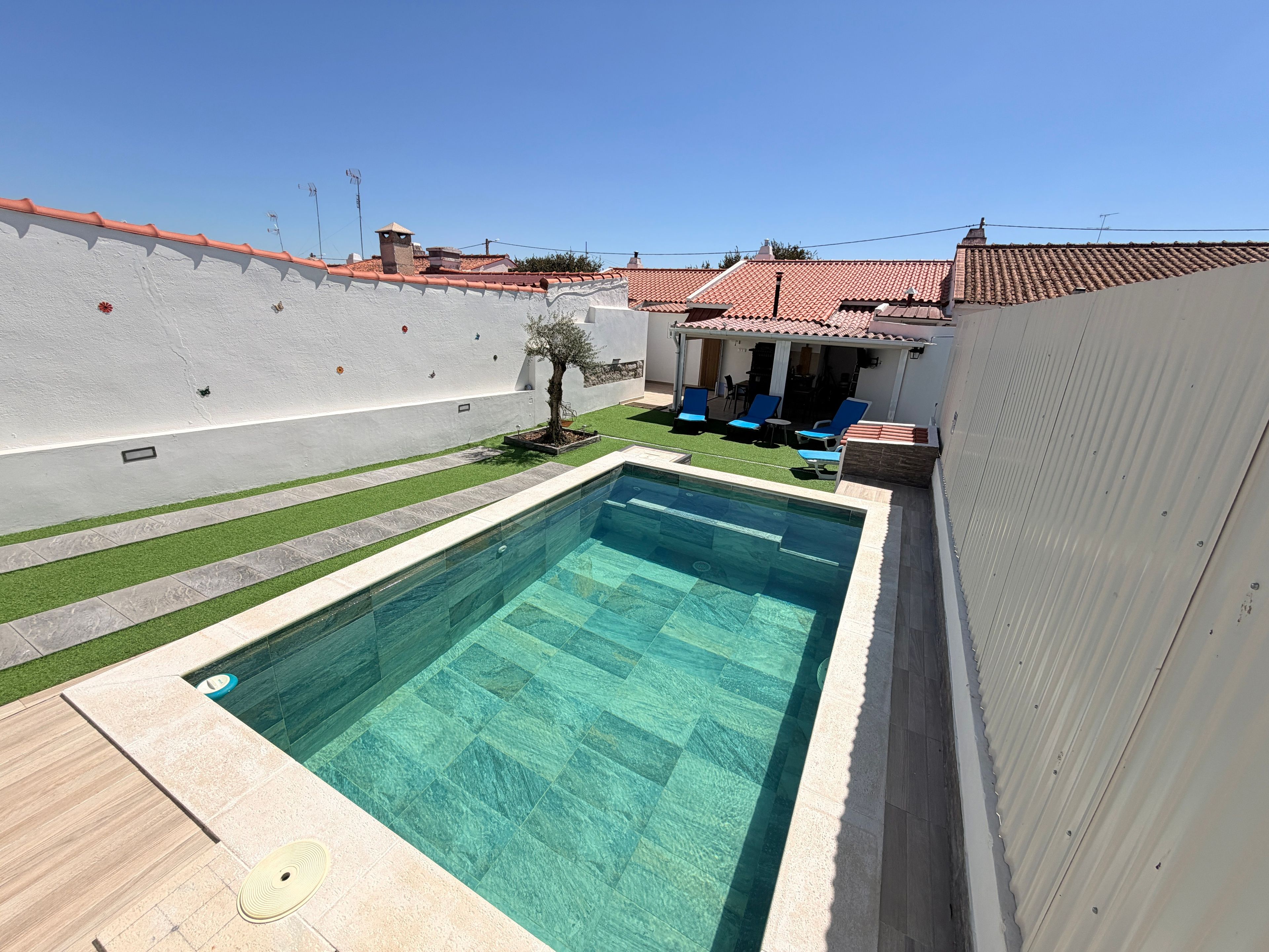 Une piscine avec une terrasse carrelée, un petit arbre et un espace patio aménagé avec des chaises.