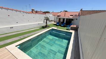 Une piscine avec une terrasse carrelée, un petit arbre et un espace patio aménagé avec des chaises.
