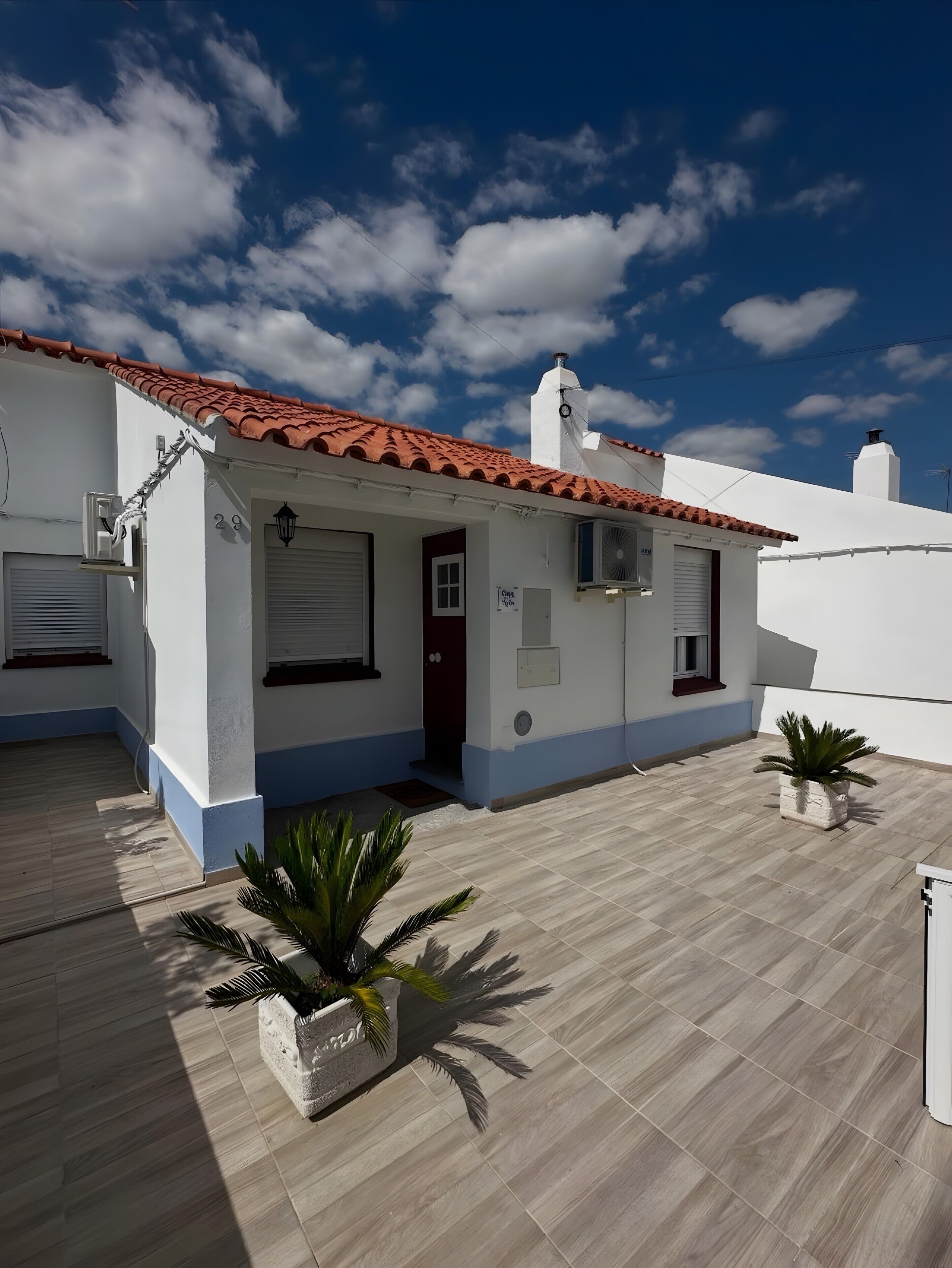 Un edificio blanco con techo de tejas rojas, una entrada pequeña y un patio con plantas en macetas.
