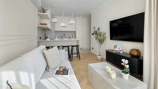 Ein modernes Wohnzimmer mit einer Couch, einem Couchtisch und einem an der Wand montierten Fernseher. Es gibt einen Küchenbereich mit einem Esstisch und Stühlen.