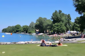Ein Park am Seeufer, auf dem sich Menschen auf der Wiese entspannen, ein Boot liegt an der Anlegestelle und im Hintergrund ist ein kleines Gebäude zu sehen.