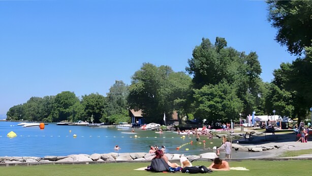 Un parc au bord du lac où des personnes se détendent sur l’herbe, un bateau amarré sur l’eau et un petit bâtiment en arrière-plan.
