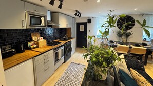Uma cozinha moderna com revestimento de azulejos pretos, eletrodomésticos de aço inoxidável e bancada de madeira. Há uma área de refeições com uma mesa redonda e cadeiras, e um relógio de parede grande.