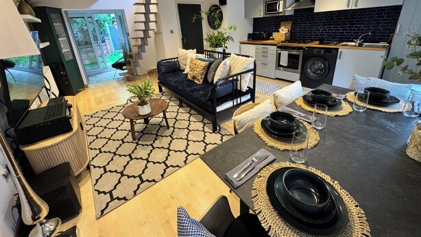 Cozinha moderna com armários preto e branco, micro-ondas e fogão. Existe uma área de jantar com mesa posta para quatro, incluindo pratos, copos e talheres. Um sofá com almofadas está posicionado perto de uma grande janela.