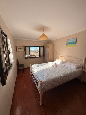 Un dormitorio con cama, ventana con vistas, una silla y un cuadro en la pared.