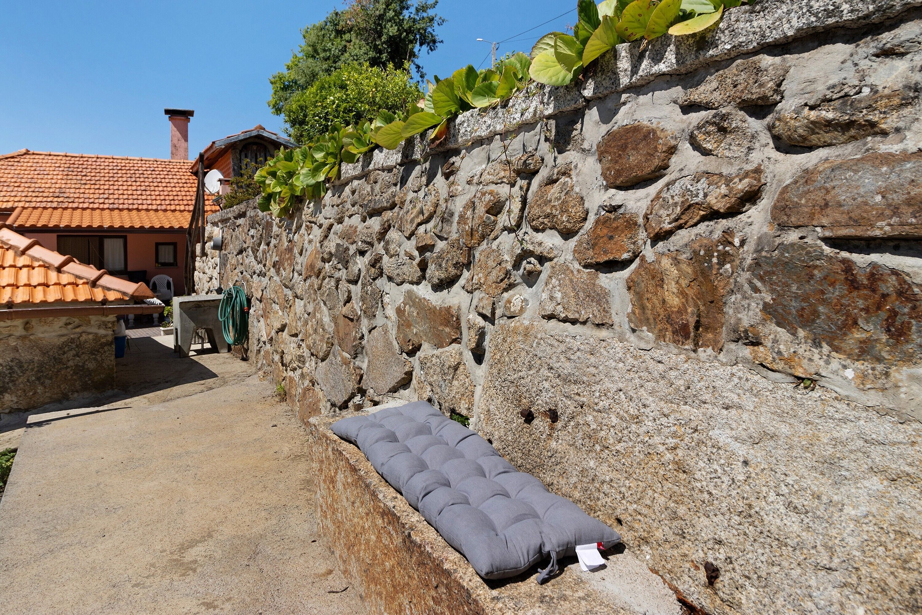 A stone wall with a mattress and a hose, a building with a tiled roof, and a clear sky.