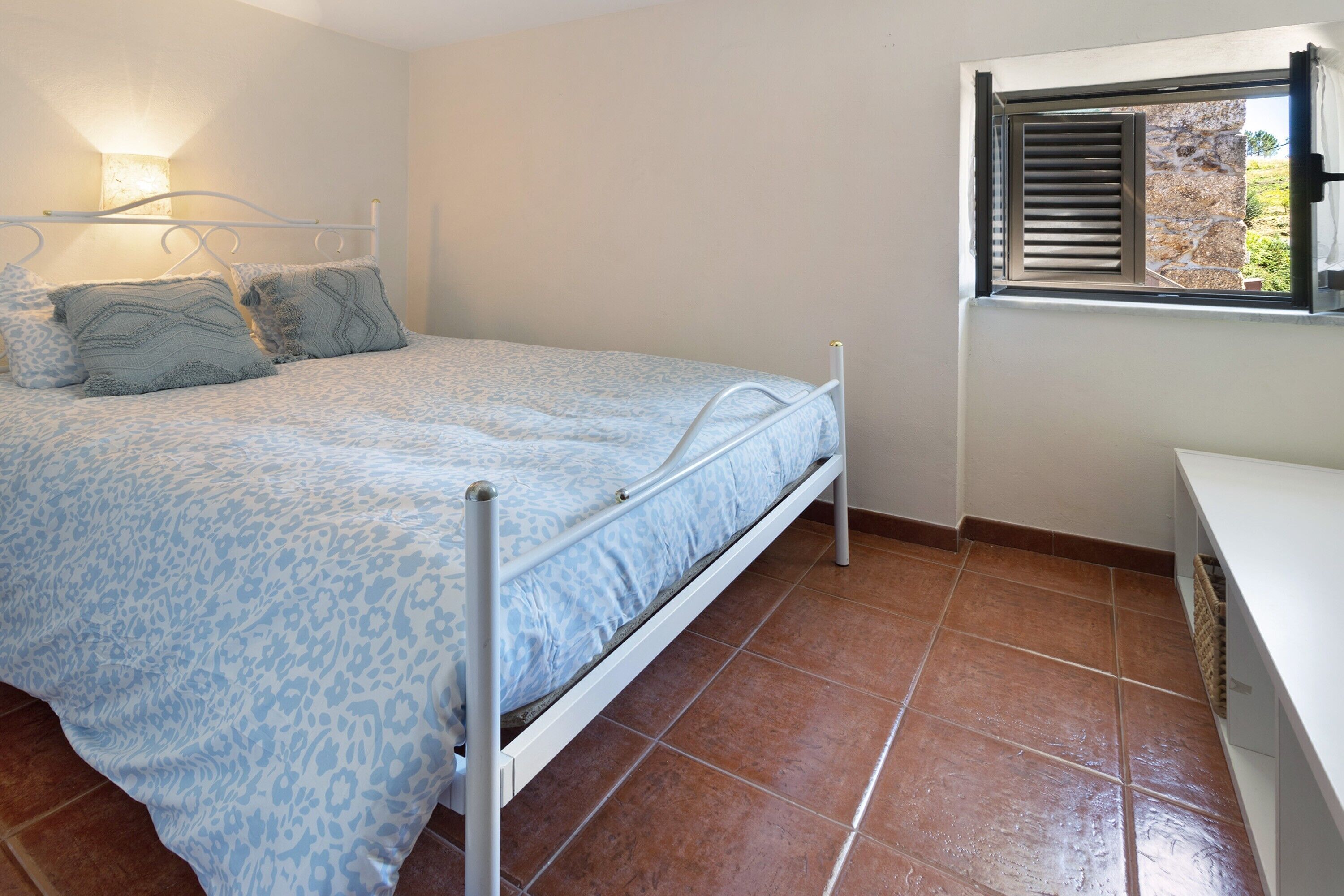 Un dormitorio con cama, almohadas, una mesita de noche, una ventana con contraventanas y una lámpara de pared.