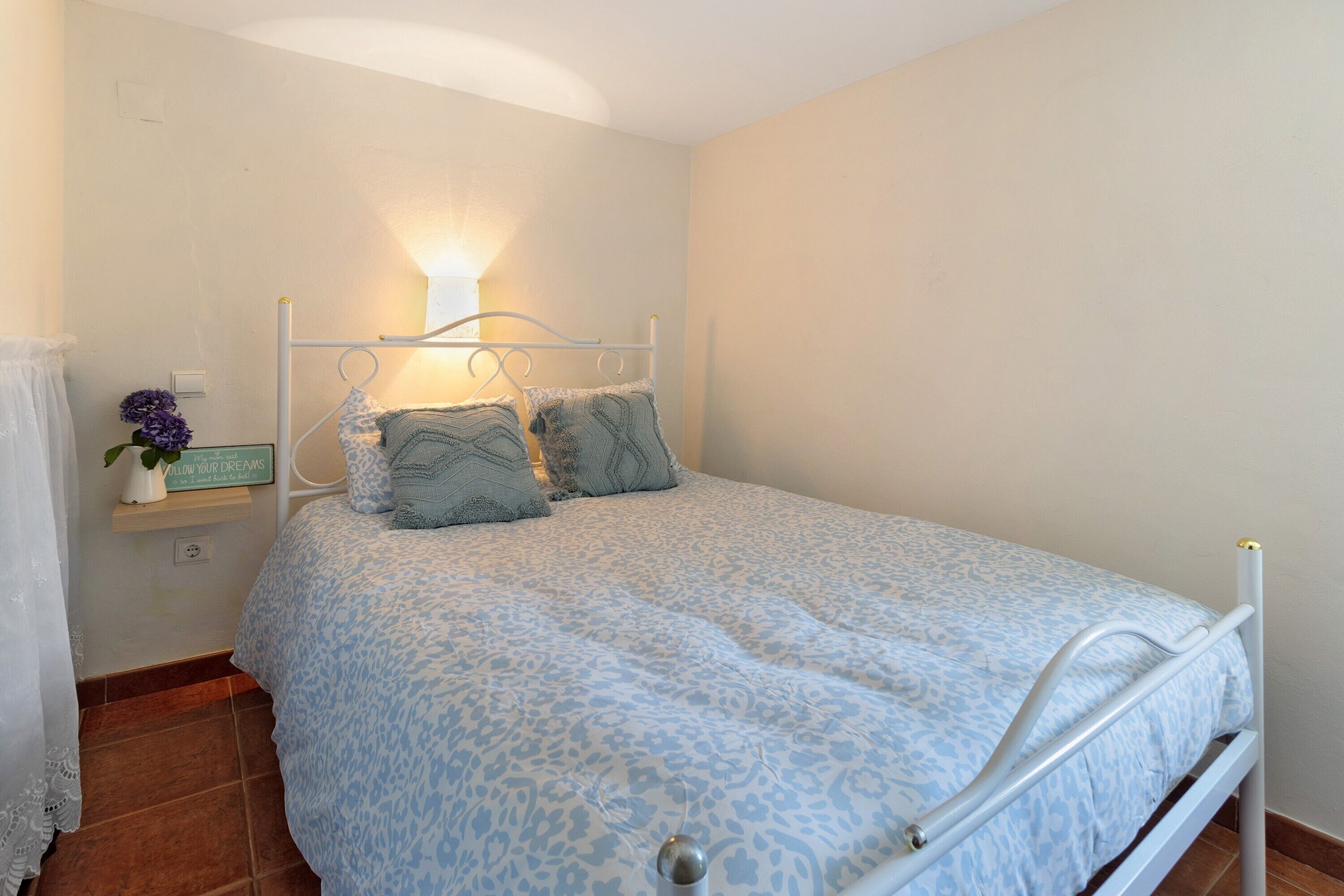 A single bed with a metal frame, a bedside table with a lamp, and a wall-mounted shelf with a potted plant.
