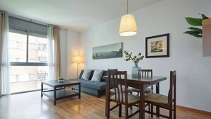 A modern living room with a sofa, dining table, chairs, a lamp, and a large window. - 5IND1114 - 1 bedroom apartment (Barcelona)